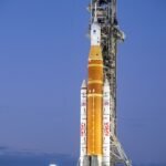 A towering orange and white NASA SLS rocket and Orion capsule stand vertically on a mobile launcher at night, brightly illuminated by spotlights against a dark sky at Launch Pad 39B.