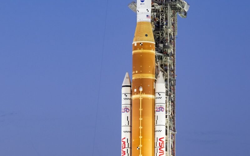 A towering orange and white NASA SLS rocket and Orion capsule stand vertically on a mobile launcher at night, brightly illuminated by spotlights against a dark sky at Launch Pad 39B.